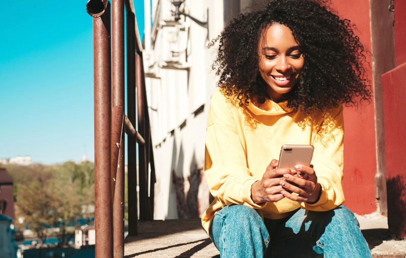 African-american-woman-searching-for-the-best-car-loan-rates-on-her-cell-phone-as-she-sits-on-stairs-wearing-a-yellow-sweatshirt.
